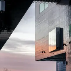 Surowa forma budynku, przypominająca brutalizm, z geometrycznymi panelami i odbijającymi światło oknami, tworzy dramatyczny kontrast z pastelowym niebem.