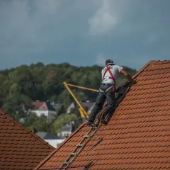 Dekarz w uprzęży bezpieczeństwa wspina się po drabinie na dach. To właśnie dekarz zajmuje się kryciem dachów.