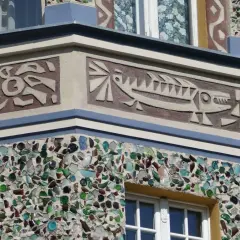 Sgraffito z rybą i abstrakcyjnymi kształtami na fasadzie budynku, ozdobionej mozaiką z kolorowego szkła.