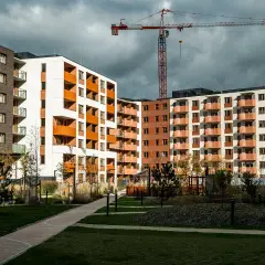 Nowe bloki mieszkalne z balkonami i żurawiem budowlanym w tle. Widać zielony dziedziniec, ścieżki i drzewa. Rynek wtórny w fazie rozwoju.