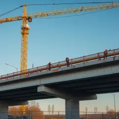 Na budowie mostu pracownicy w kaskach zastanawiają się, ile zarabia budowlaniec na godzinę. Żuraw góruje nad nimi.