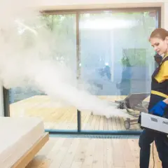 Profesjonalne ozonowanie mieszkania. Kobieta w fartuchu obsługuje generator ozonu, który rozpyla białą mgłę, odświeżając wnętrze.