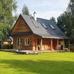 Drewniany dom kanadyjski z werandą, otoczony zielenią i drzewami. Na dachu widoczne okna dachowe i komin.