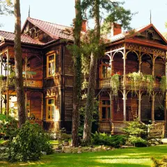 Drewniany, zabytkowy **świdermajer** z werandami i bogatymi zdobieniami, otoczony zielenią i drzewami.