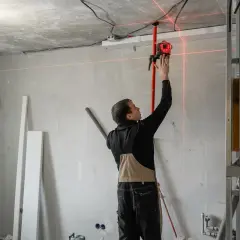 Budowlaniec ustawia laser, który wyznacza idealny pion murarski. Czerwone linie lasera przecinają pomieszczenie, pomagając w precyzyjnym montażu.