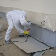 Pracownik w białym kombinezonie wykonuje malowanie elewacji pistoletem natryskowym.