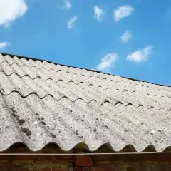 Stary dach pokryty falistym eternitem, z widocznymi plamami mchu, pod błękitnym niebem z chmurami.