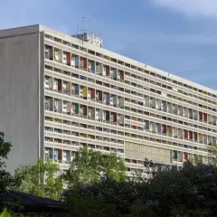 Długi blok mieszkalny w stylu modernizm architektura, z rzędami balkonów w kolorowych obramowaniach, otoczony zielenią.