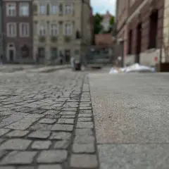 Nawierzchnia placu miejskiego, gdzie kostka brukowa sąsiaduje z betonem. W tle widać budynki i ślady prac remontowych, symbolizujące postępującą betonoza.