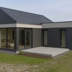 Nowoczesny dom z płyty warstwowej, z dużymi przeszkleniami i betonowym tarasem, otoczony zielenią.