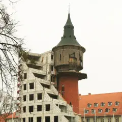 Budowa nowoczesnego budynku obok starego pylonu wieży ciśnień. Drzewa bez liści.