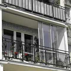 Zabudowa balkonu w bloku z przeszklonymi drzwiami i metalową balustradą. Na balkonie kwiaty w doniczkach.