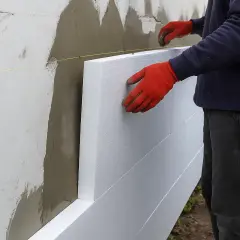 Pracownik w czerwonych rękawicach montuje płyty styropianowe na ścianie, ocieplając fasadę budynku.