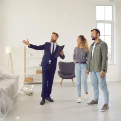 Agent prezentuje parze mieszkanie na wynajem krótkoterminowy. Wnętrze jest jasne, z meblami w folii ochronnej.