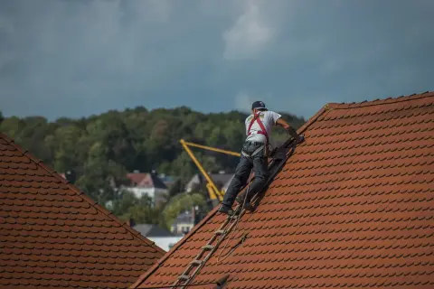 Dekarz w uprzęży bezpieczeństwa wspina się po drabinie na dach. To właśnie dekarz zajmuje się kryciem dachów.
