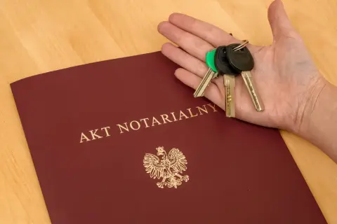 Dłoń z kluczami spoczywa na okładce dokumentu "AKT NOTARIALNY", symbolizując finalizację zakupu mieszkania.