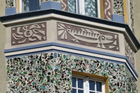 Sgraffito z rybą i abstrakcyjnymi kształtami na fasadzie budynku, ozdobionej mozaiką z kolorowego szkła.