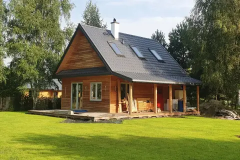 Drewniany dom kanadyjski z werandą, otoczony zielenią i drzewami. Na dachu widoczne okna dachowe i komin.