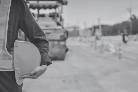 Budowa drogi, pracownik w kamizelce odblaskowej trzyma kask. W tle walec drogowy i znaki ostrzegawcze. To obraz wywłaszczenia terenu pod inwestycję.