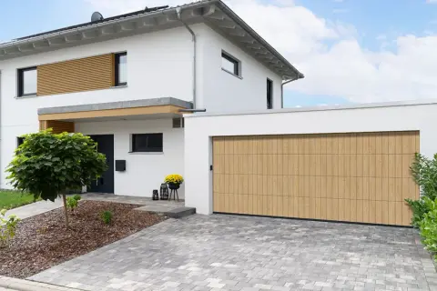 Nowoczesny dom z białym tynkiem i drewnianymi elementami. Brama garażowa w ciepłym odcieniu drewna to świetny pomysł na elewację domu, dodający mu charakteru.