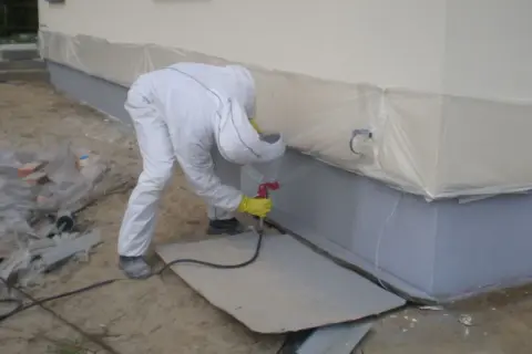 Pracownik w białym kombinezonie wykonuje malowanie elewacji pistoletem natryskowym.