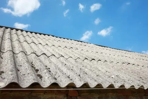Stary dach pokryty falistym eternitem, z widocznymi plamami mchu, pod błękitnym niebem z chmurami.