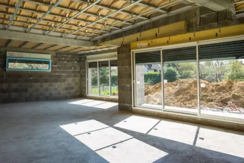 Wnętrze w budowie z betonową podłogą i ścianami. Widoczna konstrukcja sufitu i okna z widokiem na plac budowy. Powstaje nowa ściana działowa.