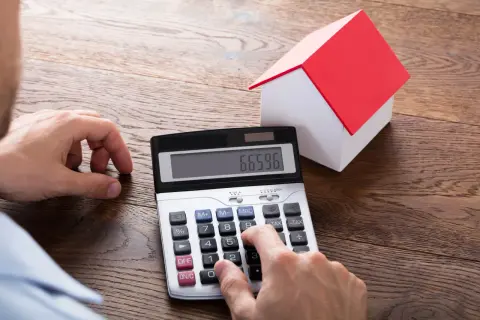 Ręce liczą na kalkulatorze, obok stoi model domu. Obliczenia dotyczą podatku od działki budowlanej.