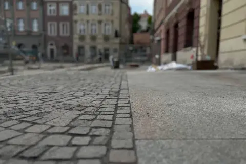Nawierzchnia placu miejskiego, gdzie kostka brukowa sąsiaduje z betonem. W tle widać budynki i ślady prac remontowych, symbolizujące postępującą betonoza.