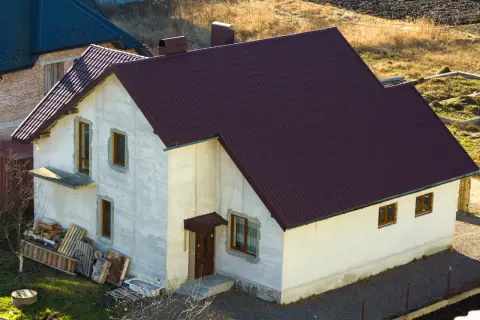 Nowy dom w zabudowie zagrodowej, z białymi ścianami i ciemnobordową dachówką. Obok materiały budowlane.