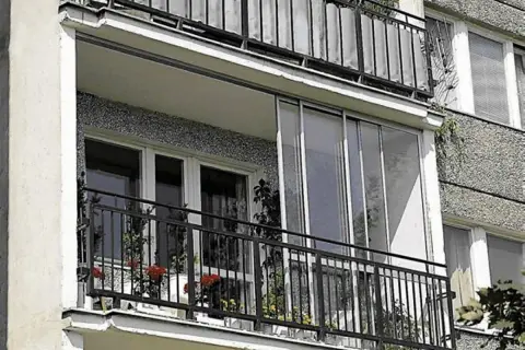 Zabudowa balkonu w bloku z przeszklonymi drzwiami i metalową balustradą. Na balkonie kwiaty w doniczkach.