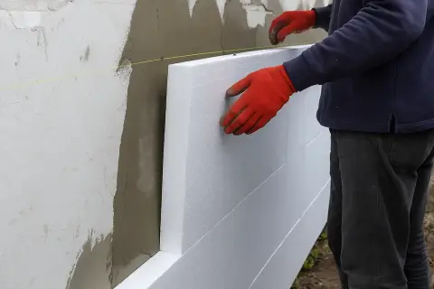 Pracownik w czerwonych rękawicach montuje płyty styropianowe na ścianie, ocieplając fasadę budynku.