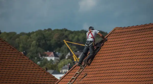 Dekarz w uprzęży bezpieczeństwa wspina się po drabinie na dach. To właśnie dekarz zajmuje się kryciem dachów.