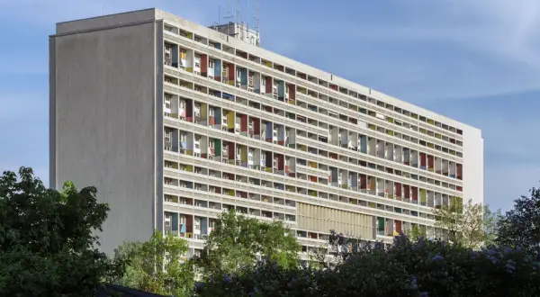 Długi blok mieszkalny w stylu modernizm architektura, z rzędami balkonów w kolorowych obramowaniach, otoczony zielenią.
