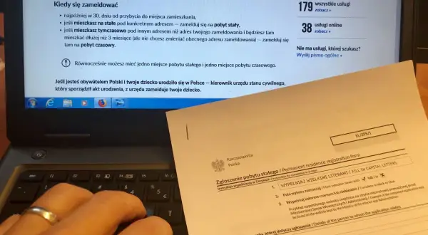Dłoń na klawiaturze laptopa, obok formularz "Zgłoszenie pobytu stałego". Obowiązek meldunkowy w praktyce.