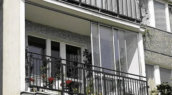 Zabudowa balkonu w bloku z przeszklonymi drzwiami i metalową balustradą. Na balkonie kwiaty w doniczkach.