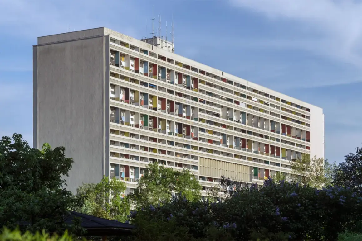 Długi blok mieszkalny w stylu modernizm architektura, z rzędami balkonów w kolorowych obramowaniach, otoczony zielenią.