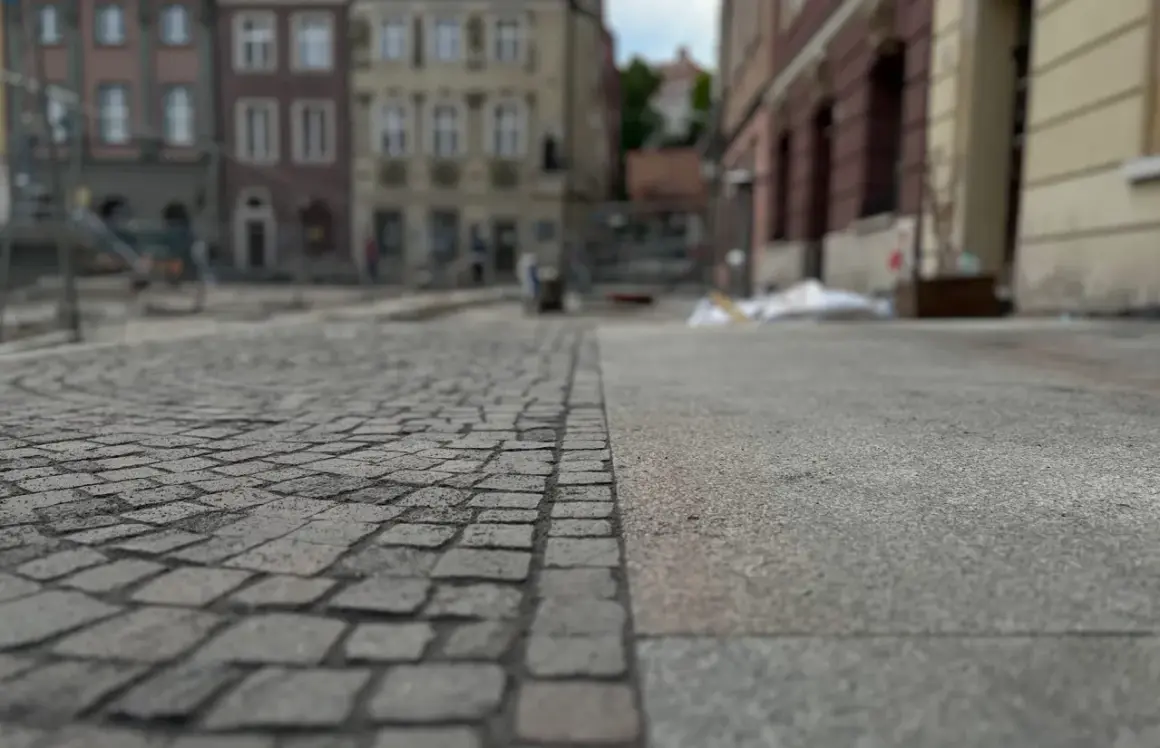Nawierzchnia placu miejskiego, gdzie kostka brukowa sąsiaduje z betonem. W tle widać budynki i ślady prac remontowych, symbolizujące postępującą betonoza.