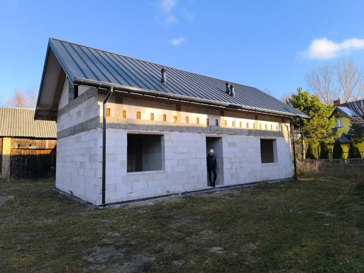 Budowa domu bez pozwolenia 70m2. Białe bloczki, szary dach, otwory okienne i drzwiowe. W tle żółty dom i drzewa.
