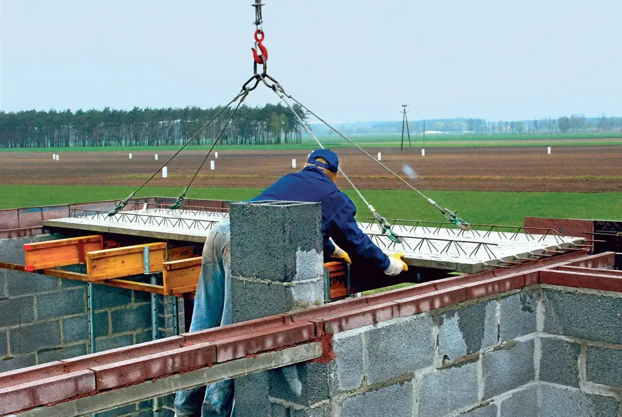 Robotnik z pomocą dźwigu montuje strop filigran. Budowa postępuje, a krajobraz wokół jest zielony.