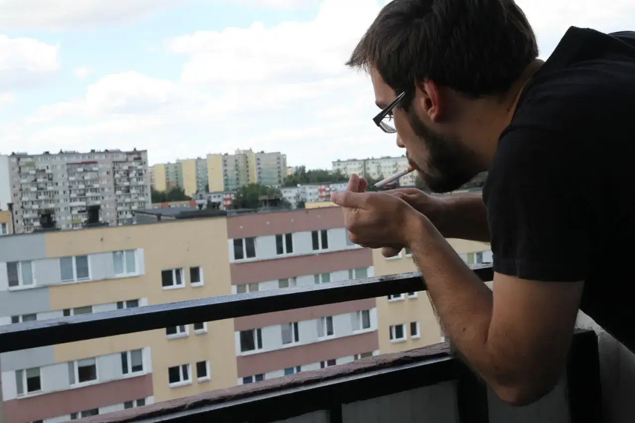 Mężczyzna zapala papierosa na balkonie, zastanawiając się, czy można palić na balkonie w bloku. W tle widać osiedle bloków mieszkalnych.