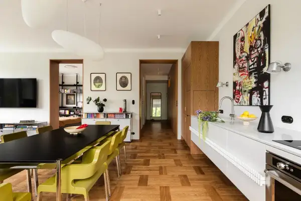 Nowoczesny salon z jadalnią i kuchnią. Czarny stół z żółtymi krzesłami, drewniana podłoga w stylu familok, kuchnia z białym blatem i drewnianymi szafkami.