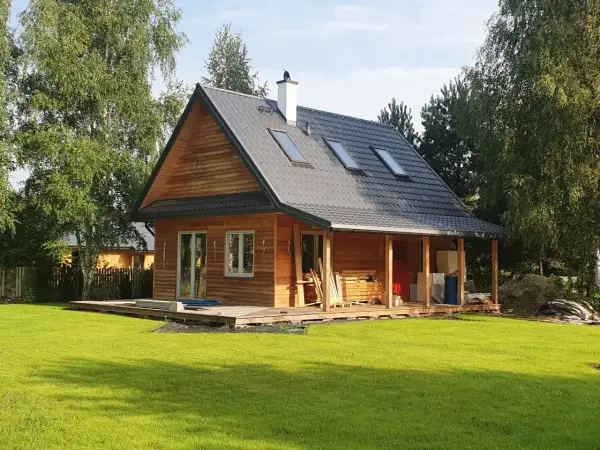 Drewniany dom kanadyjski z werandą, otoczony zielenią i drzewami. Na dachu widoczne okna dachowe i komin.