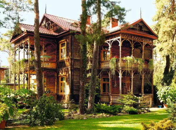 Drewniany, zabytkowy **świdermajer** z werandami i bogatymi zdobieniami, otoczony zielenią i drzewami.