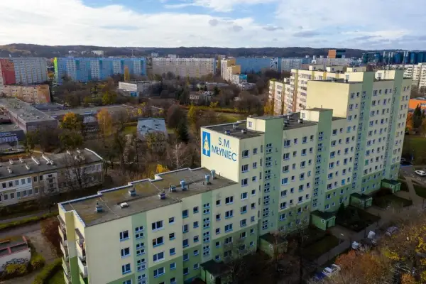 Osiedle z wielkiej płyty S.M. Młyniec, z charakterystycznymi blokami w pastelowych kolorach, otoczone jesienną zielenią i drzewami.