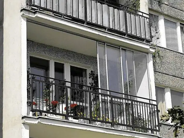 Zabudowa balkonu w bloku z przeszklonymi drzwiami i metalową balustradą. Na balkonie kwiaty w doniczkach.