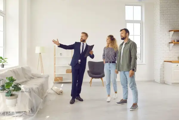 Agent prezentuje parze mieszkanie na wynajem krótkoterminowy. Wnętrze jest jasne, z meblami w folii ochronnej.